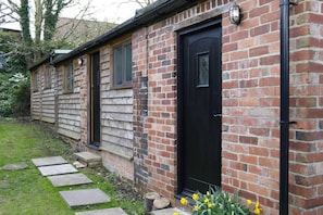 A brick building with a black door and windows, stone steps leading to the entrance, and daffodils at the base. - a Quirky Garden Building in an Orchard (Ryton)