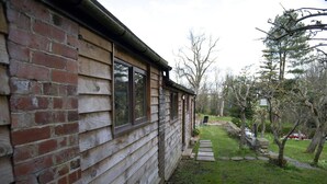 A wooden cabin with a brick foundation, a stone pathway leading to a garden, and a lawn with trees and a lawn chair. - a Quirky Garden Building in an Orchard (Ryton)