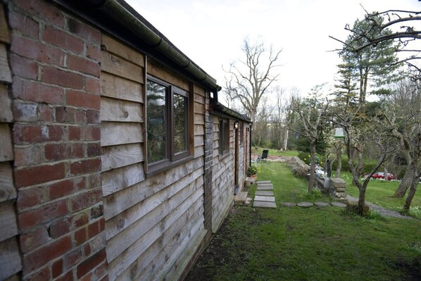 A wooden cabin with a brick foundation, a stone pathway leading to a garden, and a lawn with trees and a lawn chair.