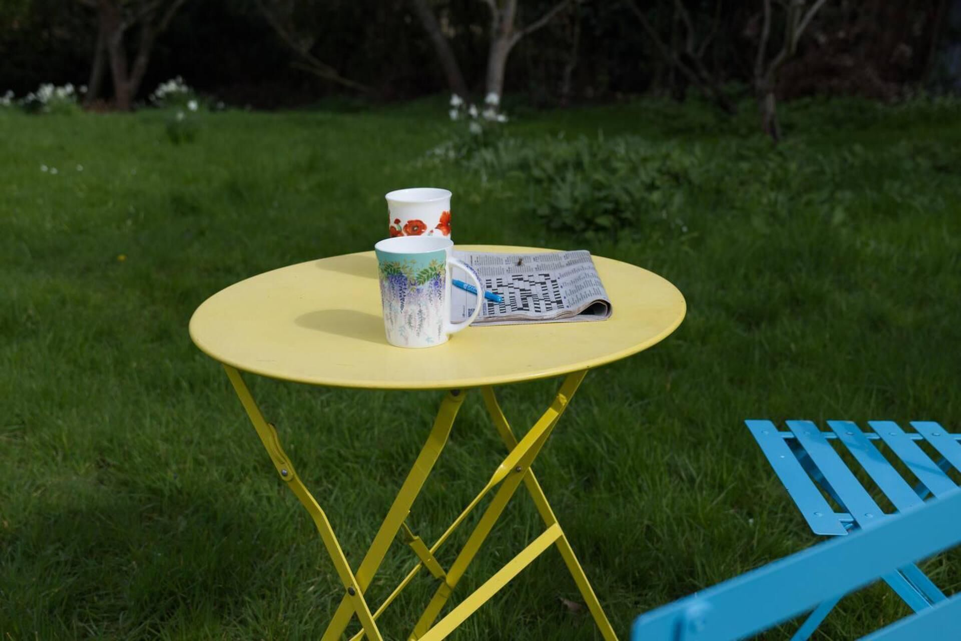 A yellow folding table with a white mug and a newspaper on it, set on grass.