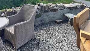 A patio area with wicker furniture and a stone wall, overlooking a scenic landscape with a bench and a fire pit. - Nuthatch - Traditional Cornish Stone Cottage (Bodmin)