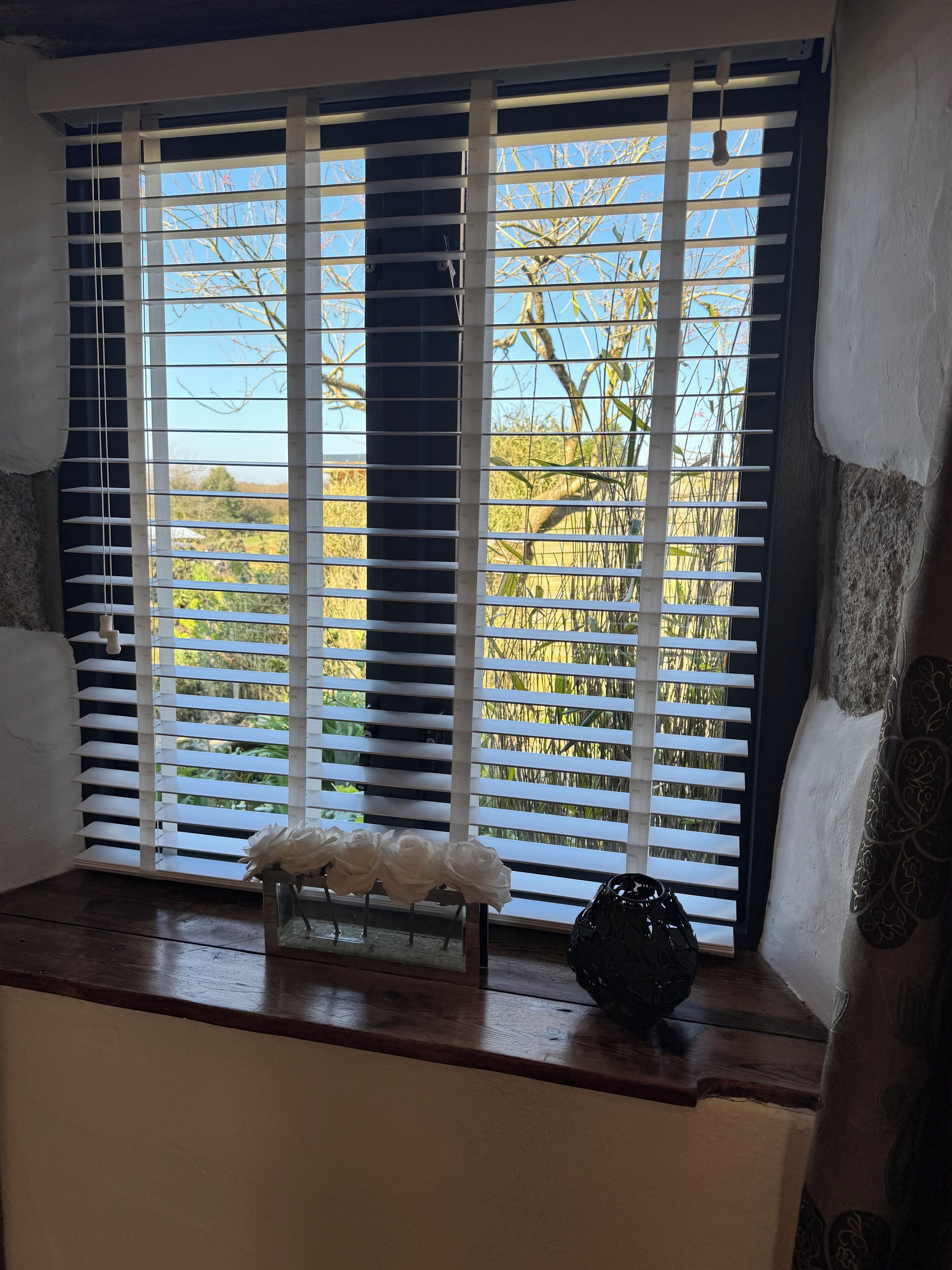 A window with white blinds, a vase with white flowers, and a decorative bowl on a wooden ledge.