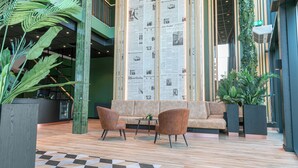 A lobby with a checkered floor, a sofa, two armchairs, a reception desk, and a large vertical display of newspapers. - Distrikt Hotels Amsterdam Zaandam  (Zaandam)