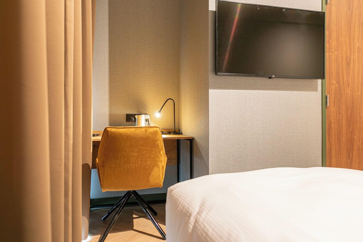 A hotel room with a bed, a desk with a chair, a TV mounted on the wall, and a lamp on the desk.
