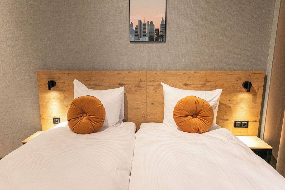 A double bed with white linens and two orange pillows, a wooden headboard, and a cityscape painting above.