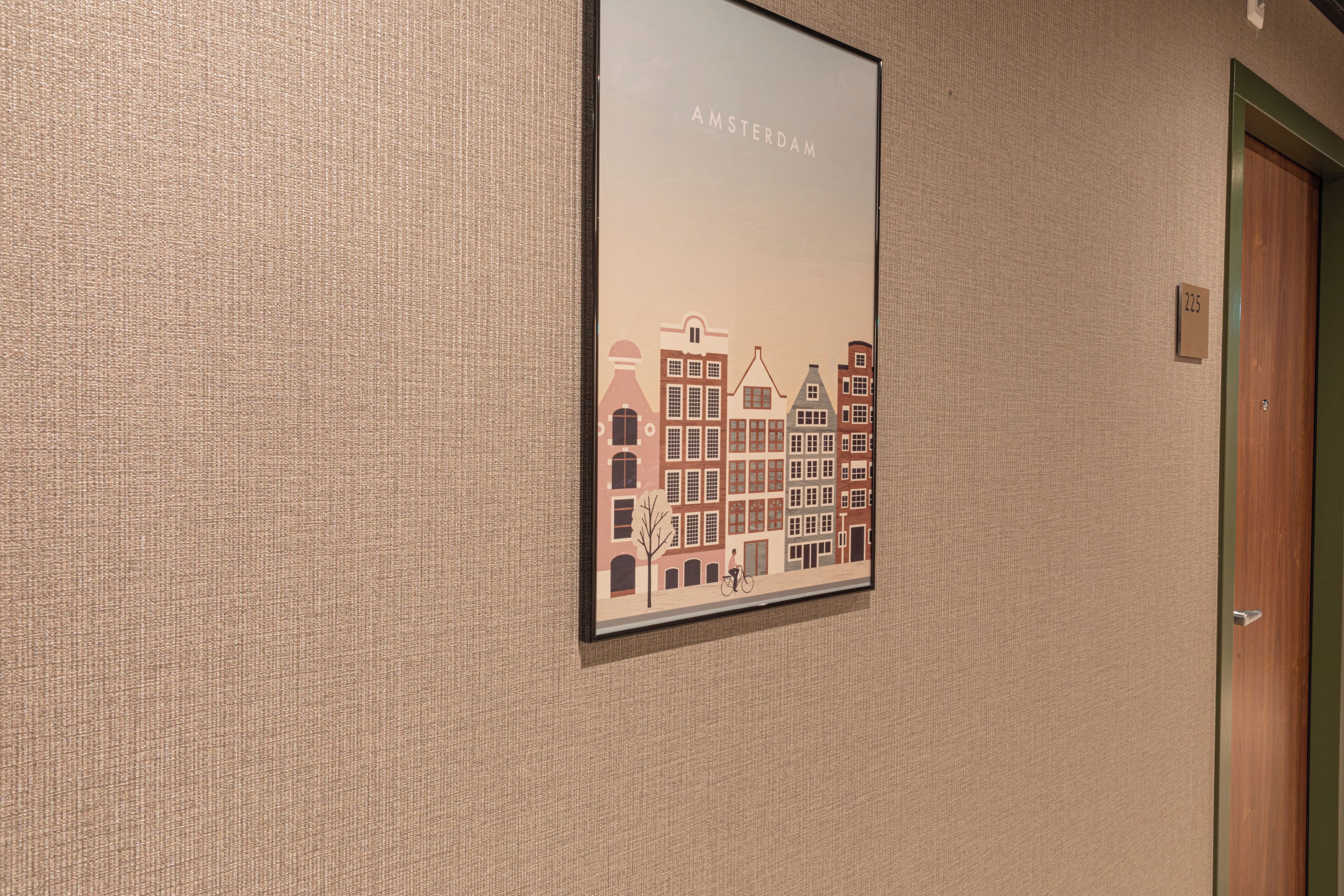 Une affiche représentant un paysage urbain, encadrée et accrochée à un mur texturé, une porte avec une plaque numérotée et un petit panneau au-dessus de la porte.
