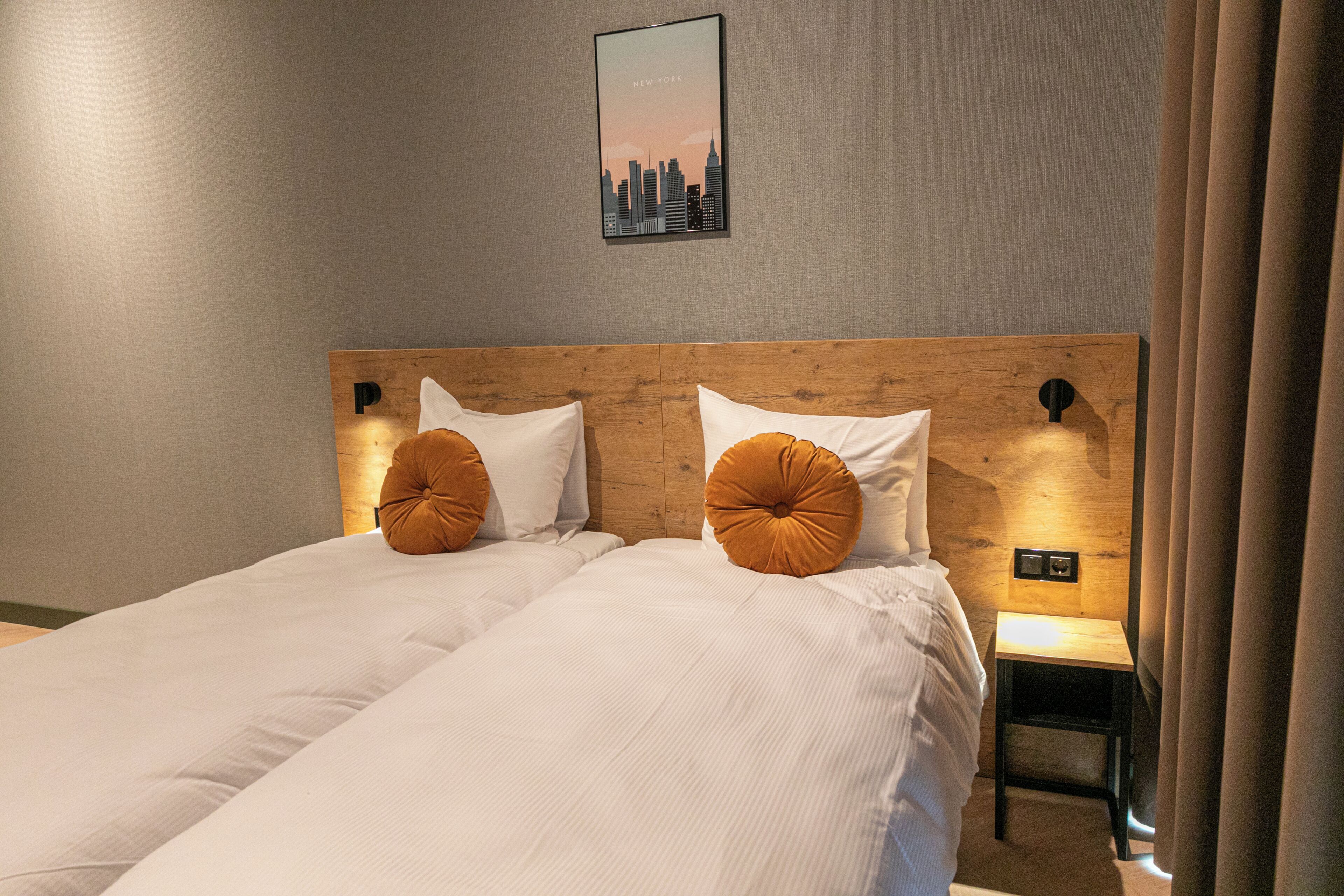 A double bed with two pillows, a wooden headboard, a nightstand with a lamp, and a cityscape poster on the wall.