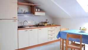 A compact kitchen with white cabinets, a sink, and a stove. There is a dining table with a blue tablecloth and wooden chairs. - Residence Belvedere (Ascea)
