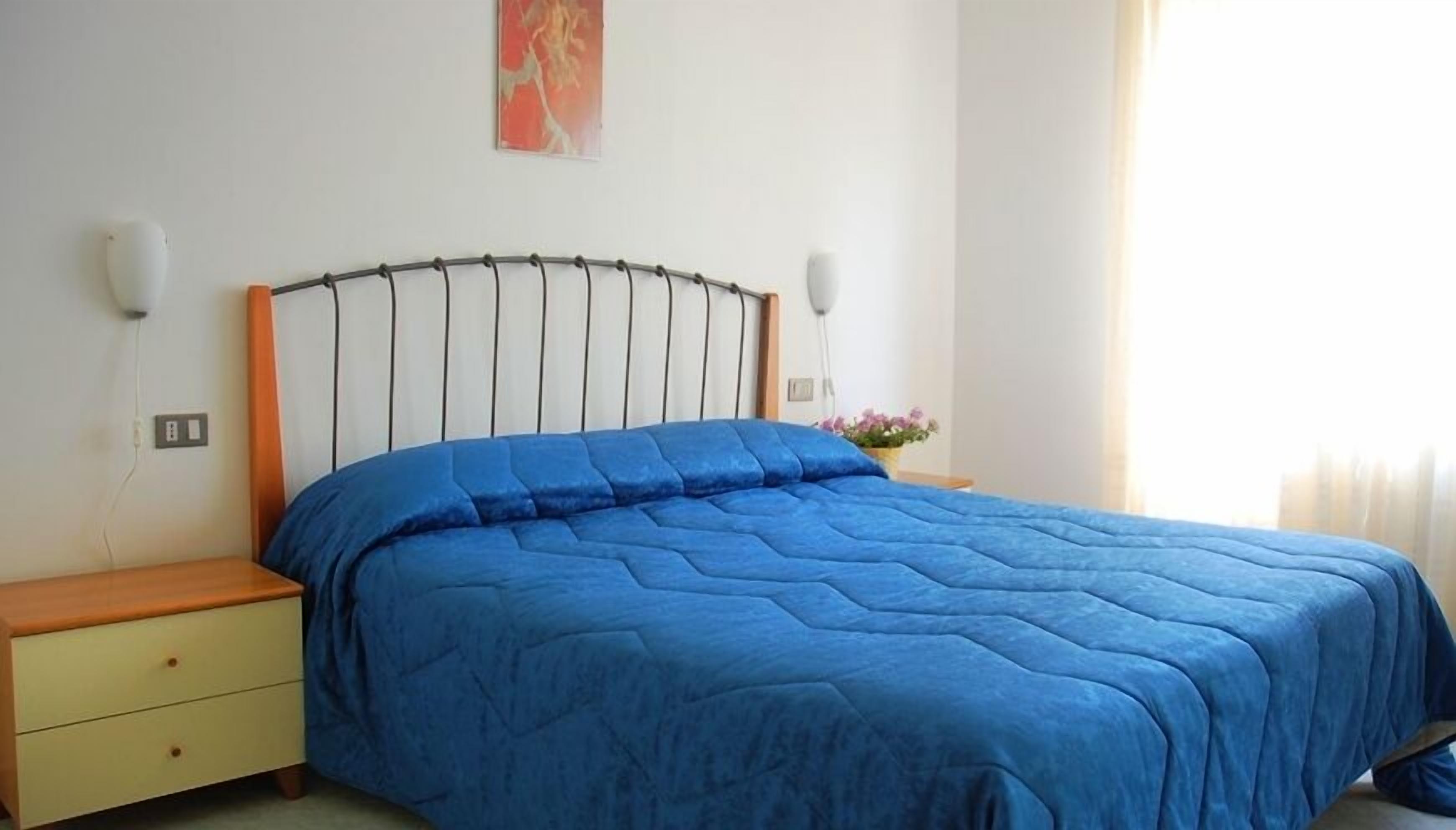Un dormitorio con cama, mesita de noche, cajón, luz empotrada en la pared y un cuadro.