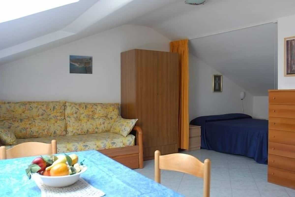 A small, cozy room with a wooden wardrobe, a blue bed, a floral sofa, and a dining table with fruit.