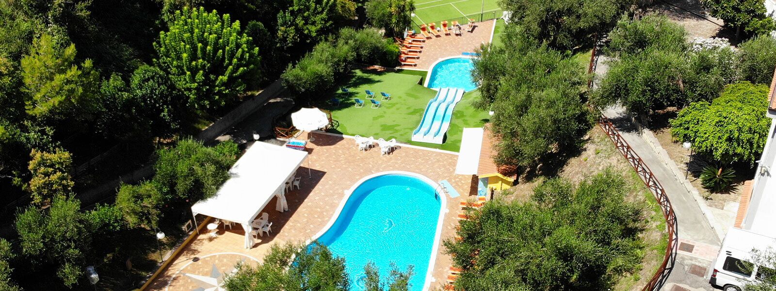 Vista aérea de um resort com uma grande piscina, um tobogã aquático e uma quadra de tênis.