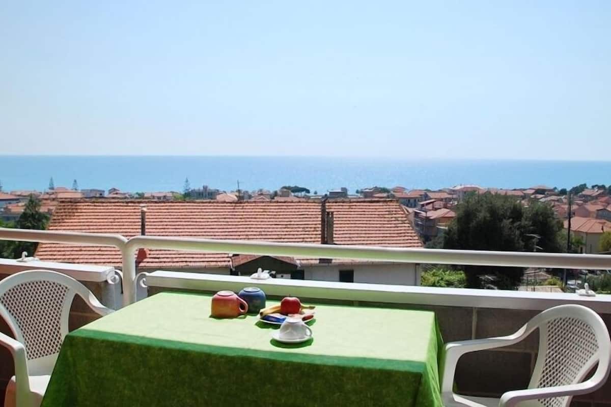A balcony with a table set for tea, overlooking a coastal town with terracotta rooftops.
