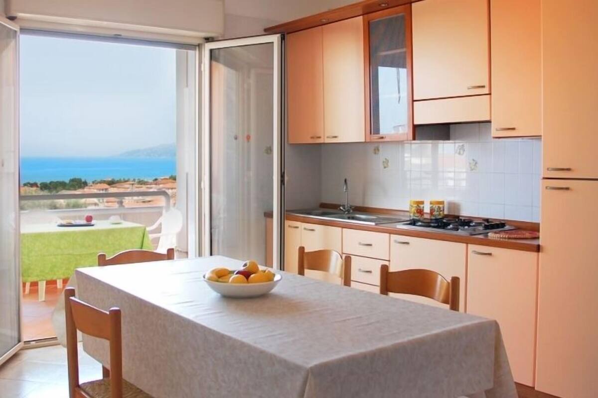 A kitchen with a view of the sea, a dining table with fruit, and a balcony with a table and chairs.