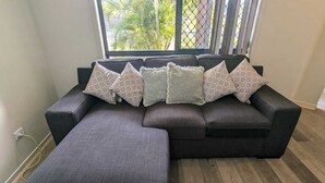 A sectional sofa with a matching ottoman in a room with wooden flooring and a window with a view of palm trees. - Gladstone Super Home Fully Fenced With Central Aircondioning (New Auckland)