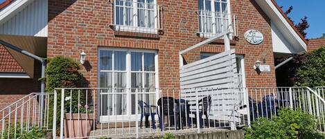 Una casa de ladrillo de dos pisos con fachada blanca, un balcón con barandilla blanca y un pequeño jardín delantero.