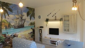 A living room with a mural of a ship on a beach, a TV mounted on the wall, and a dining table with chairs. - Il Veliero Appartamento  con Giardino (Terracina)