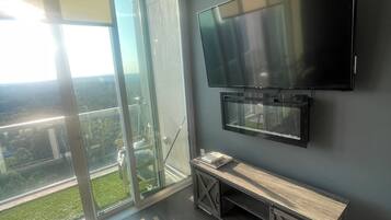 A modern living room with a large flat-screen TV mounted on a dark wall, a wooden bench, and a sliding glass door leading to a balcony with a view of greenery.