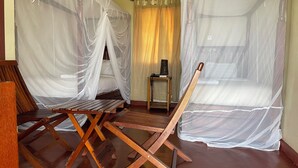 Quarto com uma cama coberta por mosquiteiro branco, uma cadeira de madeira, uma mesinha com abajur e uma cortina.