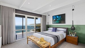A bedroom with a large window overlooking the sea, a bed with a striped blanket, a bedside table with a lamp, and a painting on the wall.