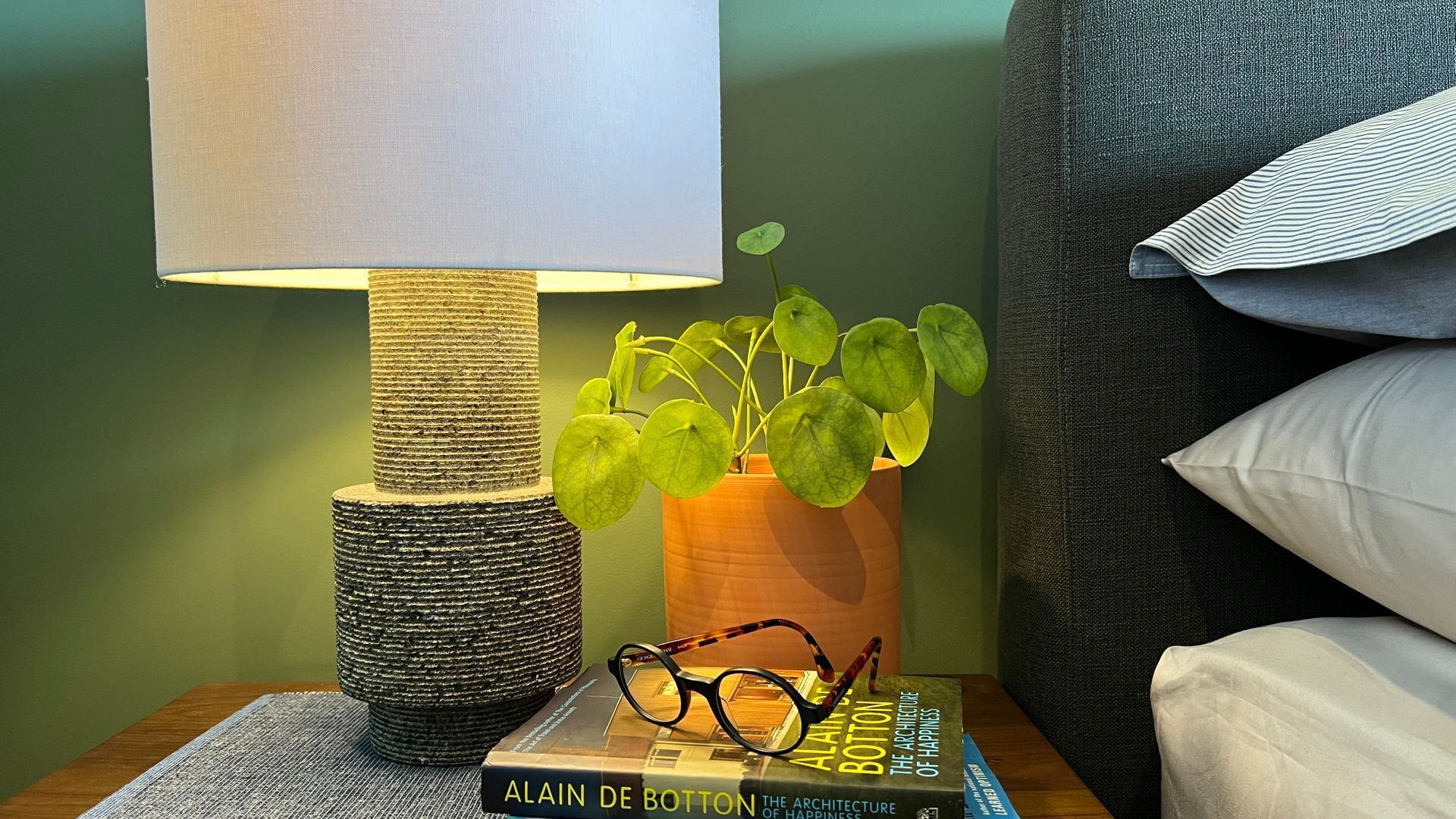 Un comodino con una lampada, una pianta, un paio di occhiali e un libro intitolato "The Architecture of Happiness" di Alain De Botton.