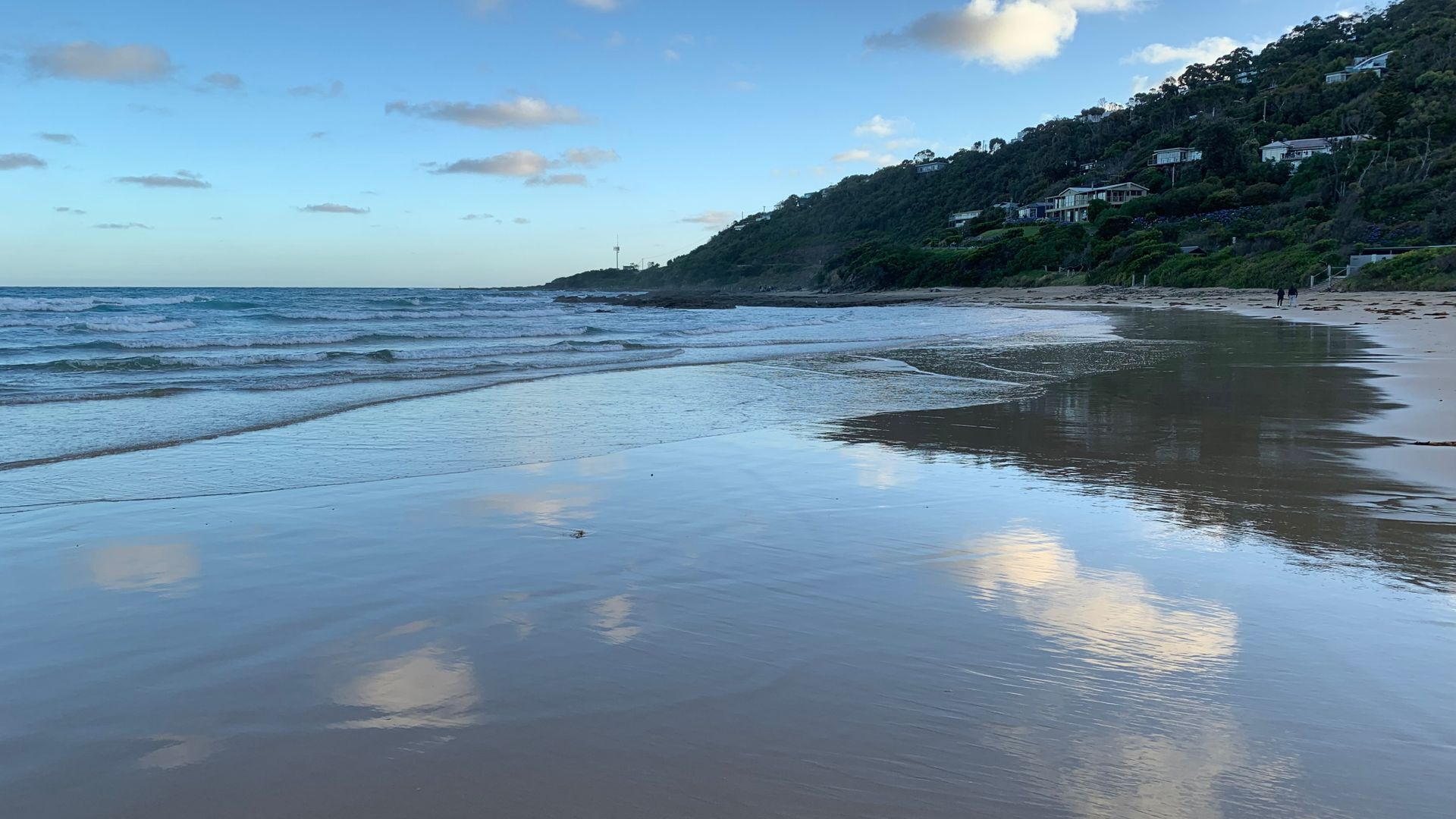 A beach with calm waves, a clear sky, and houses on a hillside in the background.