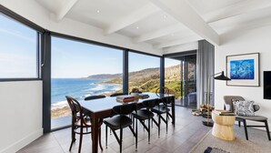 A modern dining area with a large table and chairs, large windows offering a view of the ocean and hills, and a cozy seating area with a lamp and cushions.