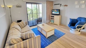 Uma sala de estar com um sofá, uma mesa de centro, uma TV sobre um suporte, um tapete azul e um ventilador de teto.