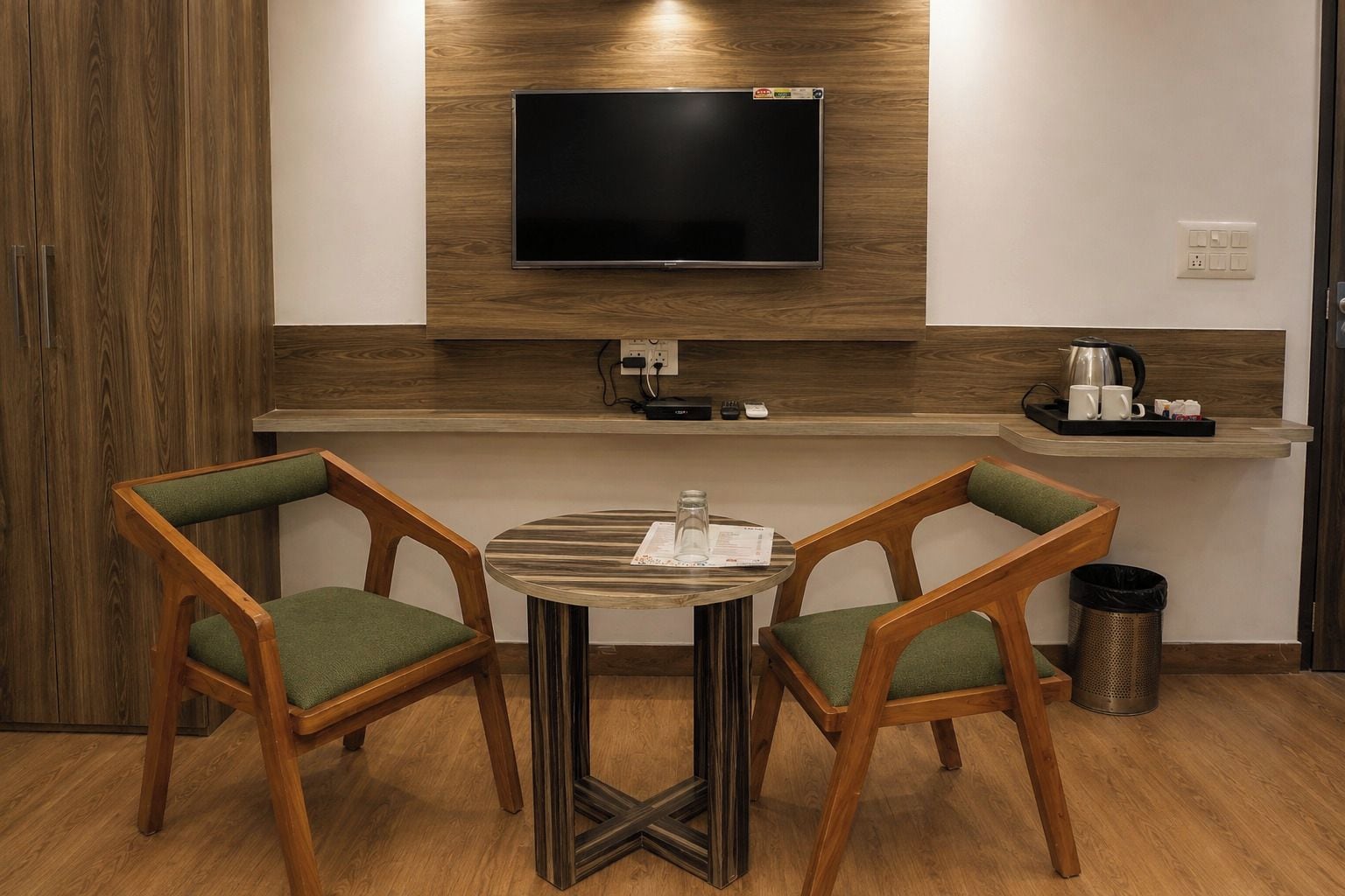 A small, modern room with a wooden floor, a round table with two chairs, a flat-screen TV mounted on the wall, a small shelf with a kettle and cups, and a trash can.