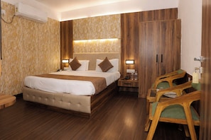 Quarto de hotel com cama grande, guarda-roupa de madeira, mesas de cabeceira e um espaço de estar com cadeiras.
