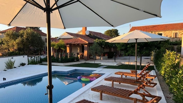 Un espace piscine doté de chaises longues et de parasols, entouré d’un jardin et de bâtiments.