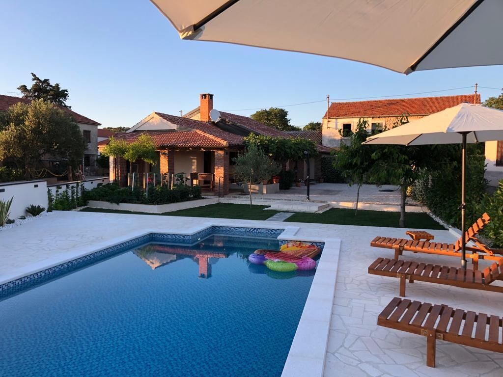 A swimming pool with lounge chairs and umbrellas, surrounded by a garden and a house with a tiled roof.