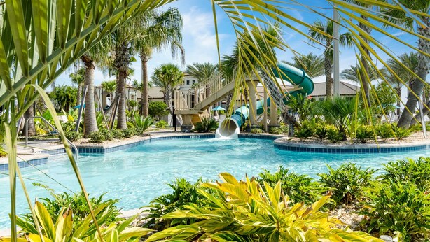 A swimming pool with a water slide, surrounded by palm trees and a building with a covered walkway. - "Enchanted Escape" - Game Room|Massage Chairs|Peloton Bike|Sleeps 23 (Davenport)