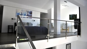 Un vaste espace de vie doté d’une balustrade en verre, d’un escalier et offrant une vue sur une chambre à coucher avec un lit et un tableau.