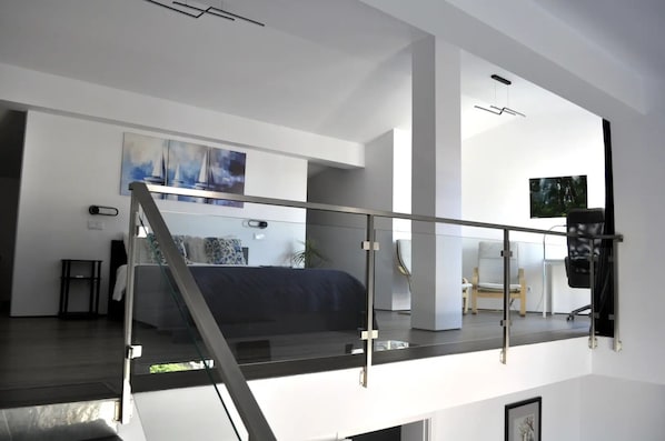 A spacious living area with a glass railing, a staircase, and a view of a bedroom with a bed and a painting.