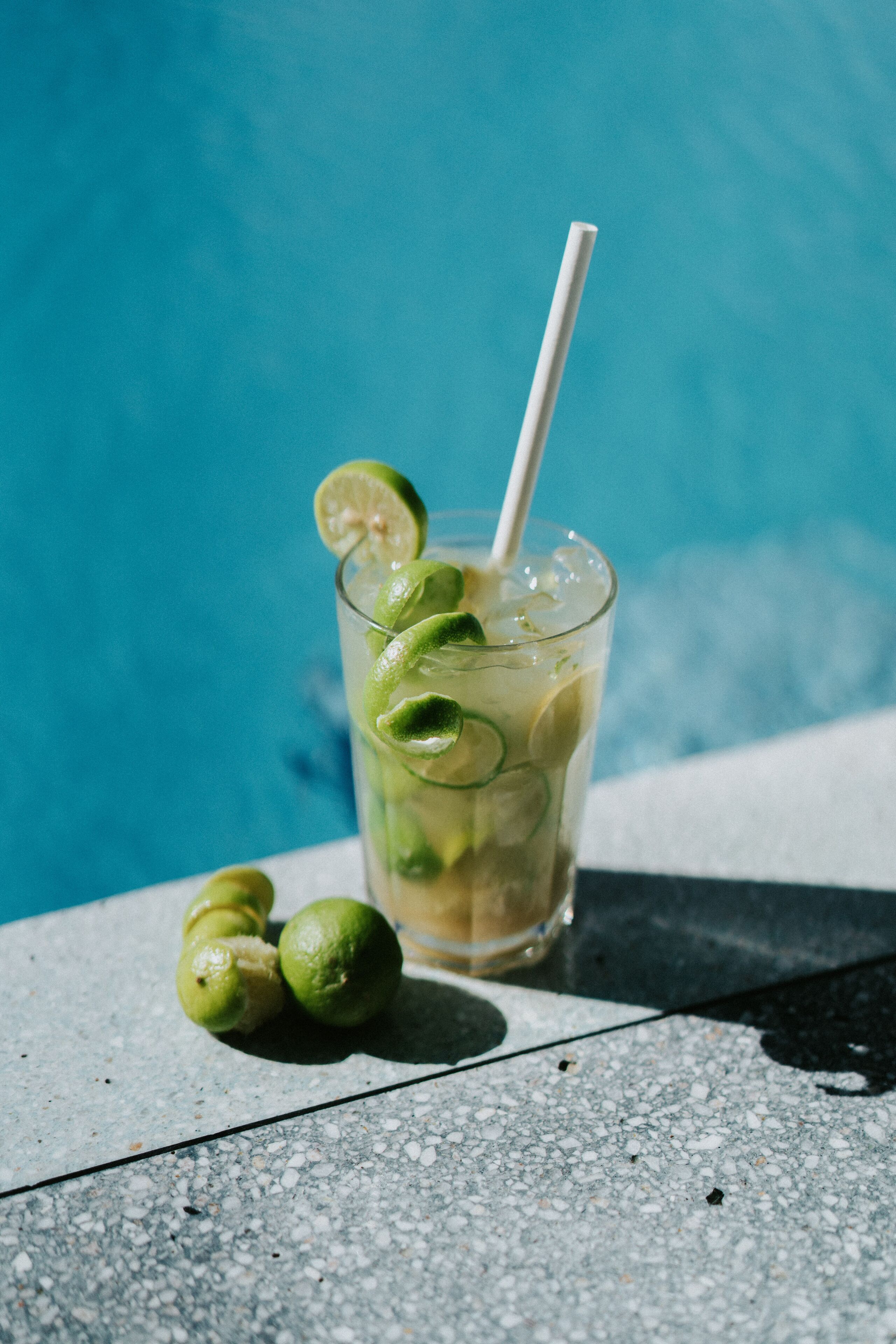 A glass of iced drink with lime slices and a straw, placed on a textured surface near a pool.