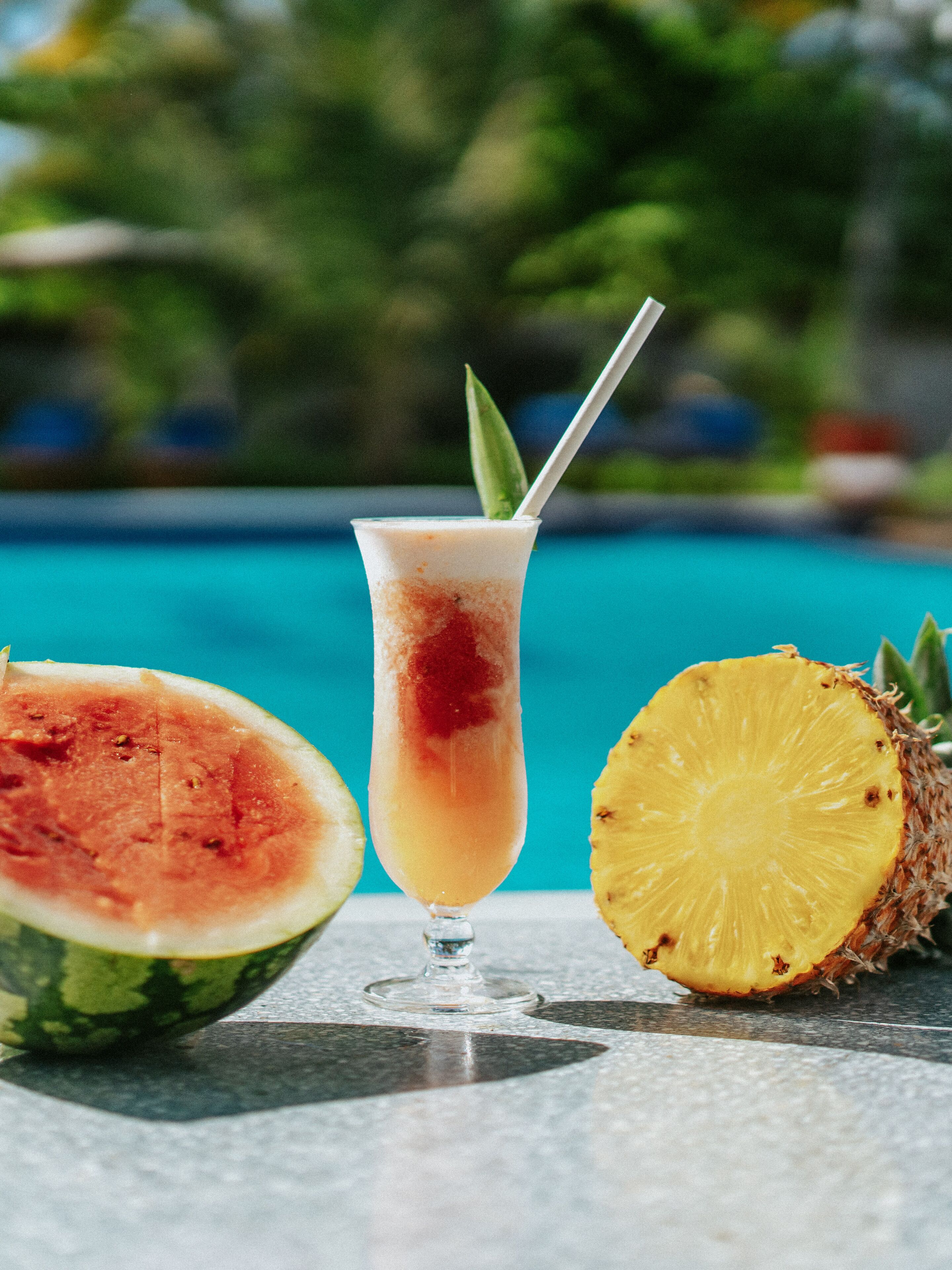 Ein Glas tropischer Drink mit Trinkhalm und einer Wassermelonen Scheibe daneben, auf einem Tisch am Pool platziert.