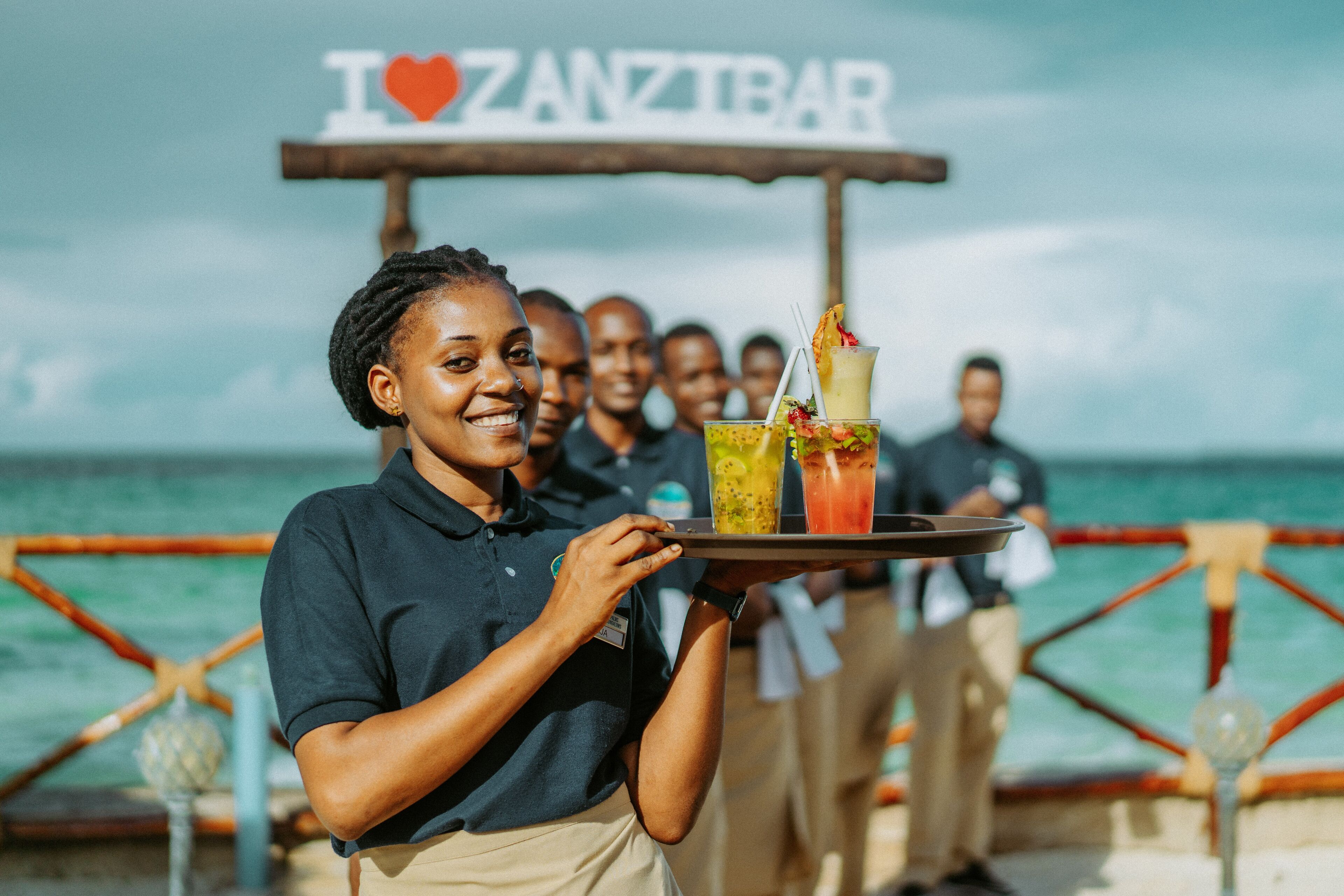 A person holding a tray with drinks at a waterfront bar with a sign that reads "I ♥ ZANZIBAR".