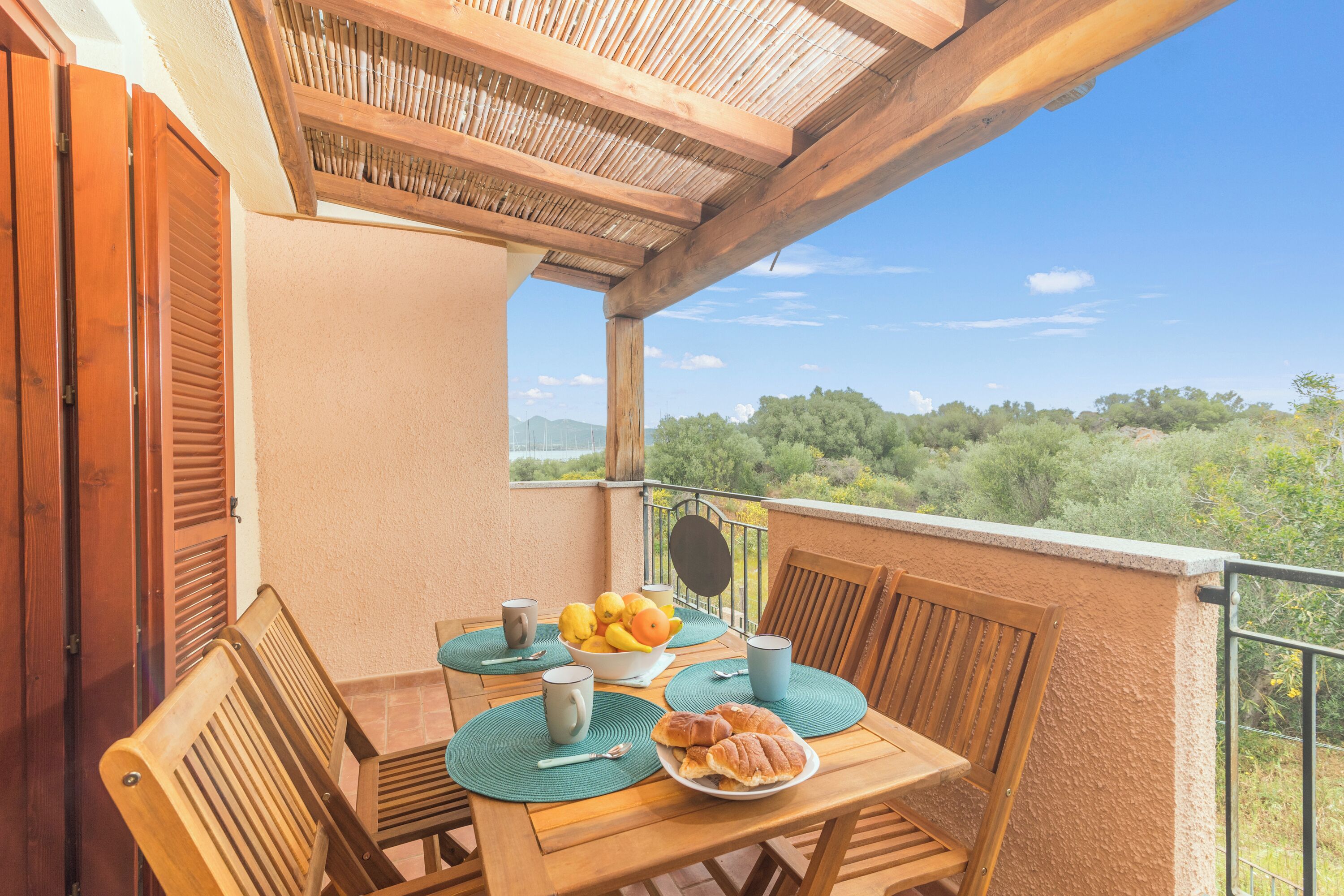 Een houten tafel met een fruitschaal, een bord met croissants en twee mokken op een balkon met uitzicht op bomen en bergen.