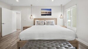 A modern bedroom with a large bed, two bedside tables, a bench, a ceiling fan, and a window with a view. - Luxury Beachfront Villa - Penthouse B (Carlsbad)