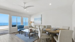 A spacious living room with a large window overlooking the ocean, a ceiling fan, a round dining table with chairs, and a sofa. - Luxury Beachfront Villa - Penthouse B (Carlsbad)
