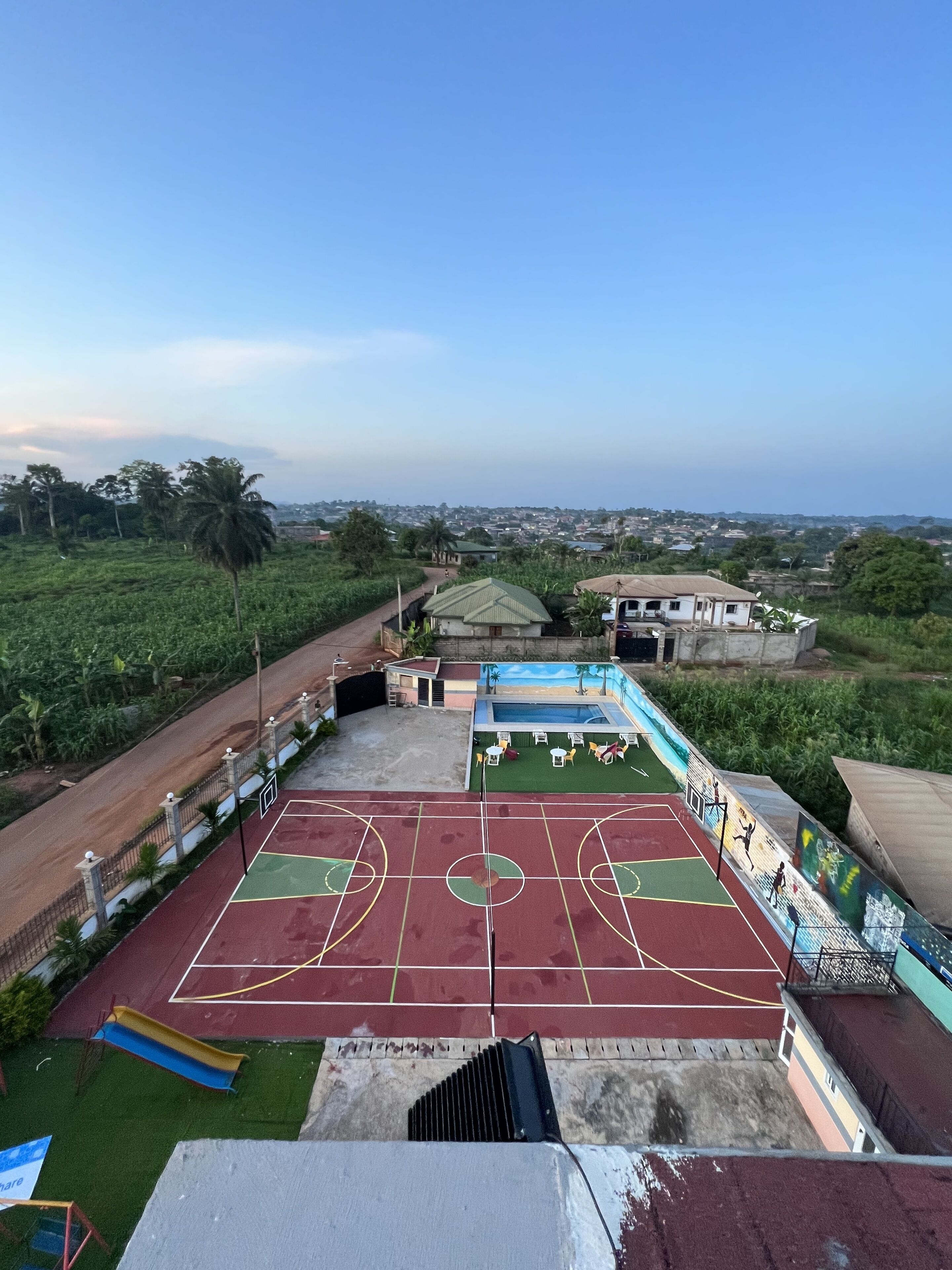 Una cancha de baloncesto al aire libre con césped verde, un edificio pequeño y vistas a un pueblo distante.