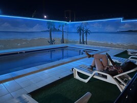 Un área de piscina con un mural al estilo playa, sillas de descanso y un suelo de baldosas.