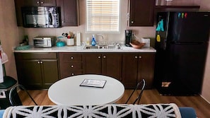 A compact kitchen with a microwave, toaster, sink, and refrigerator. There's a dining table with a placemat and a window with blinds.