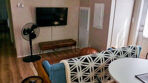 A living room with a flat-screen TV mounted on the wall, a standing fan, a bench, a patterned throw over a chair, and a small round table. - Private, detached 1 bedroom apt. 6 miles from Folly Beach & downtown Charleston (Charleston)