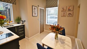 Une cuisine moderne avec un plan de travail blanc, des meubles noirs et un coin repas comprenant une table blanche et des chaises bleues. Une fenêtre est agrémentée de rideaux à motifs, et un vase de fleurs est posé sur la table.