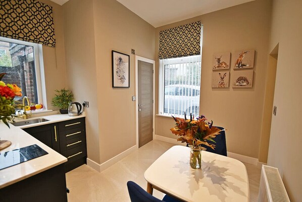A modern kitchen with a white countertop, black cabinets, and a dining area with a white table and blue chairs. There is a window with patterned curtains and a vase of flowers on the table.