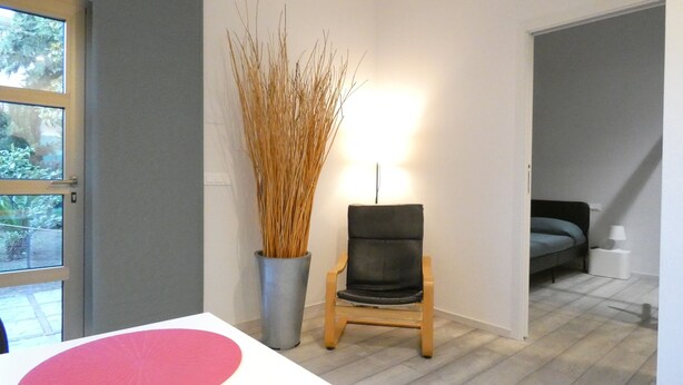 A room with a wooden chair, a tall vase with dried plants, a bed with a blue mattress, and a white bedside table. - BuenRetiroPe_Ita - Comfortable two-room apartments with garden (Pescara)
