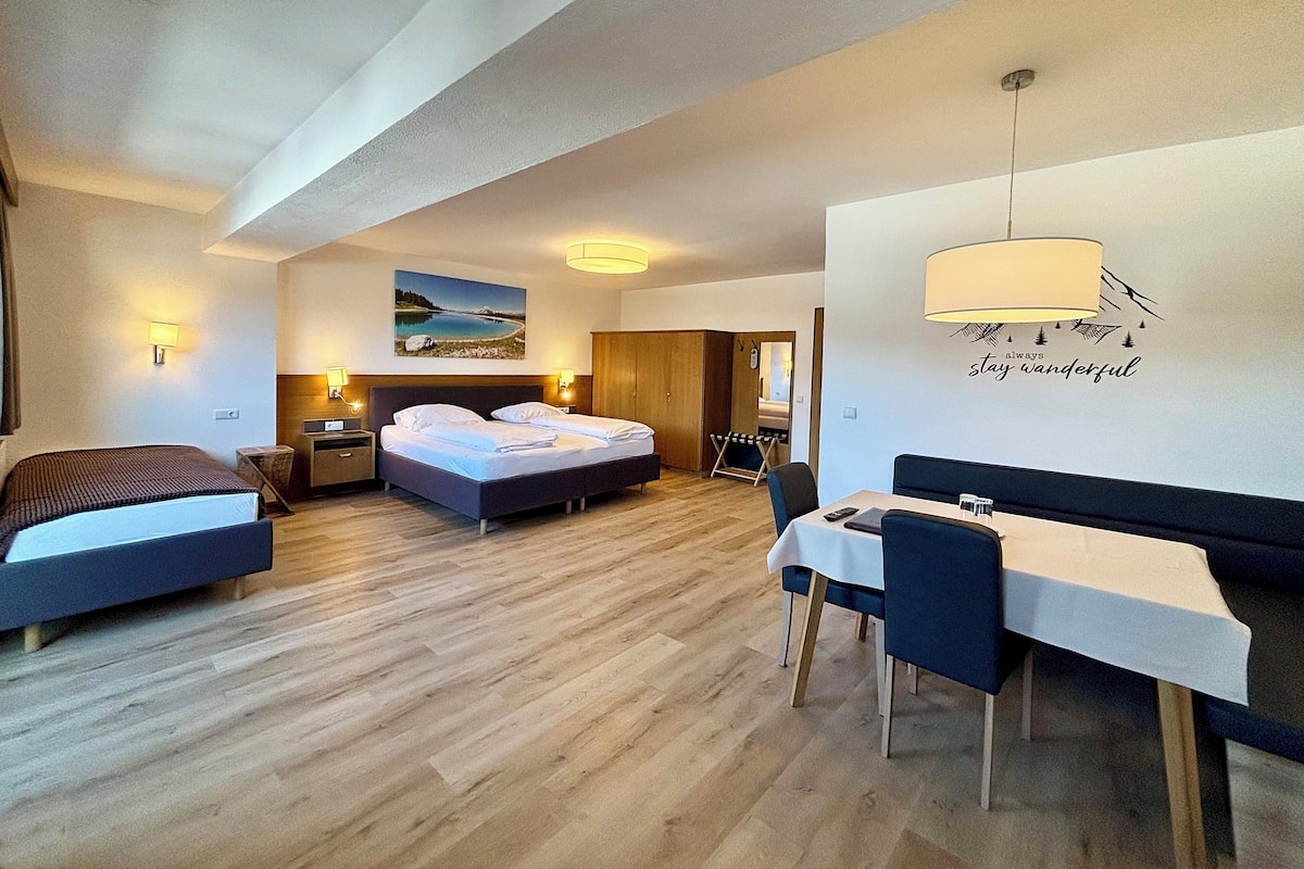 A hotel room with two beds, a desk, and a dining table. There is a painting on the wall and a sign that says "always stay wonderful."