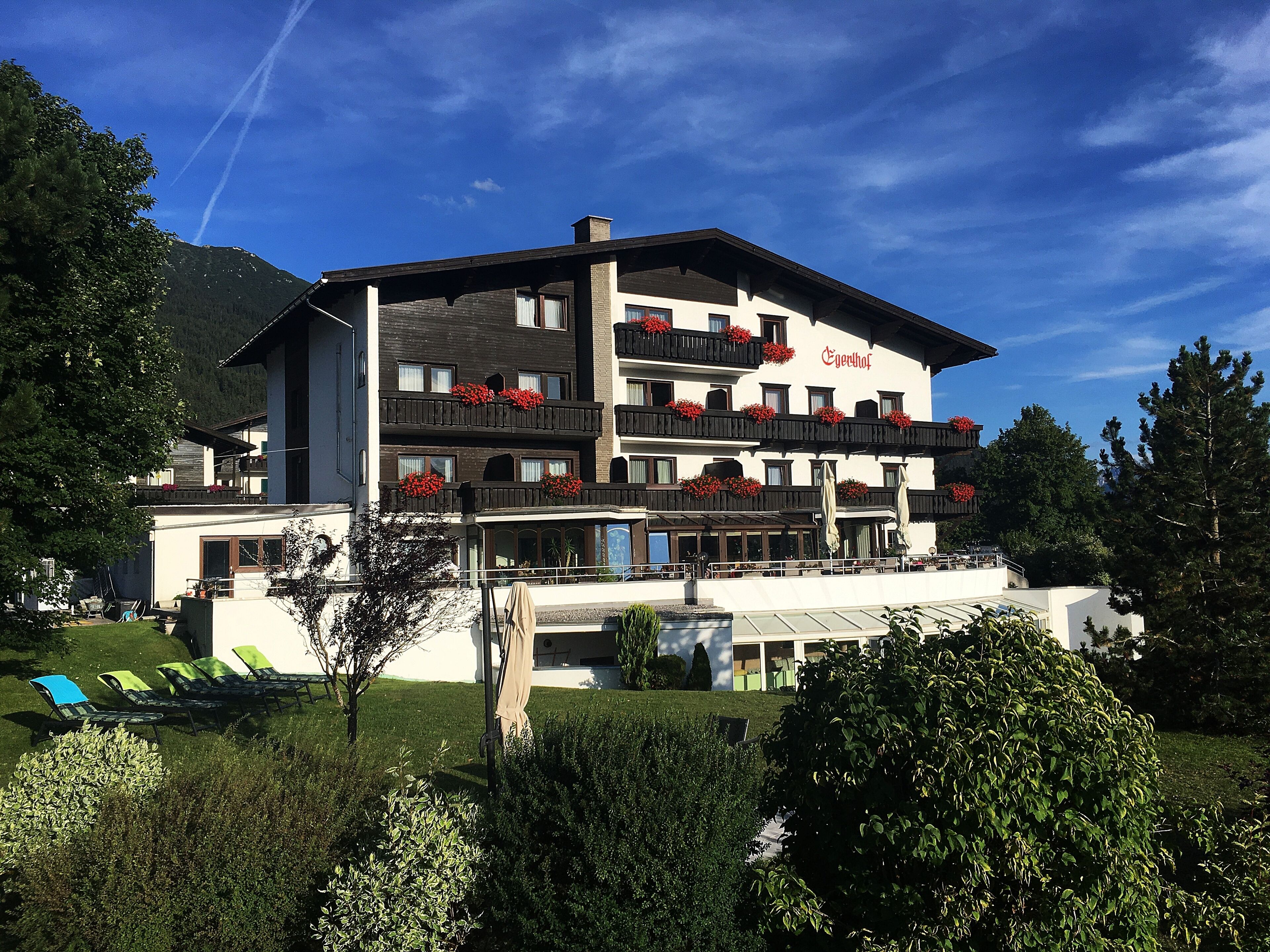 A multi-story hotel with balconies adorned with red flowers, a terrace with sun loungers, and a mountain backdrop.