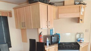 Eine kompakte Küchenzeile mit Herd, Mikrowelle und kleinem Kühlschrank. Ein Esstisch mit Stühlen und einem Mülleimer stehen bereit.